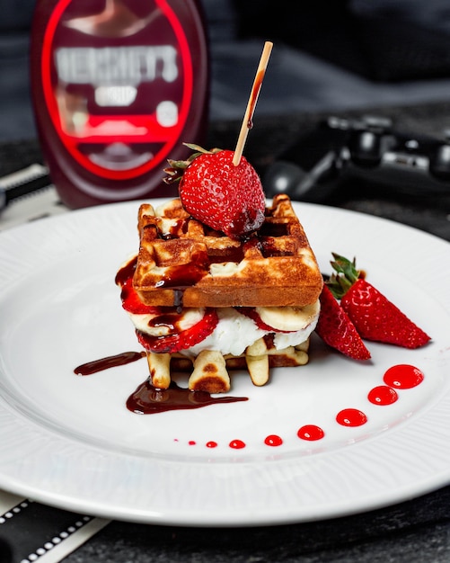 Vista lateral de waffle con helado de fresas y plátanos cubiertos con salsa de chocolate en un plato blanco