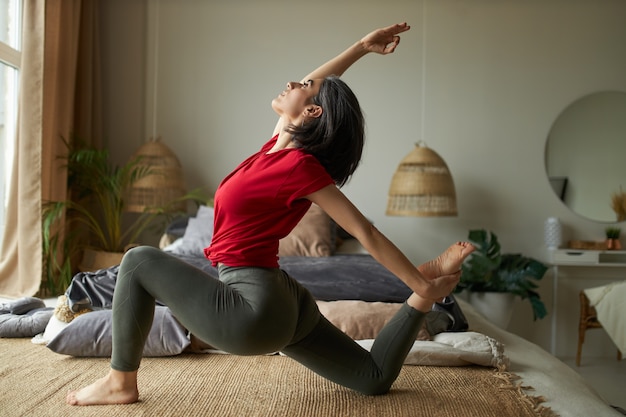Vista lateral del yogui avanzado femenino joven flexible que se ejercita en el interior haciendo Eka Pada Rajakapotasana Pose o One-Legged King Pigeon Posture II, estirando la parte frontal del torso, los tobillos, los muslos y las ingles