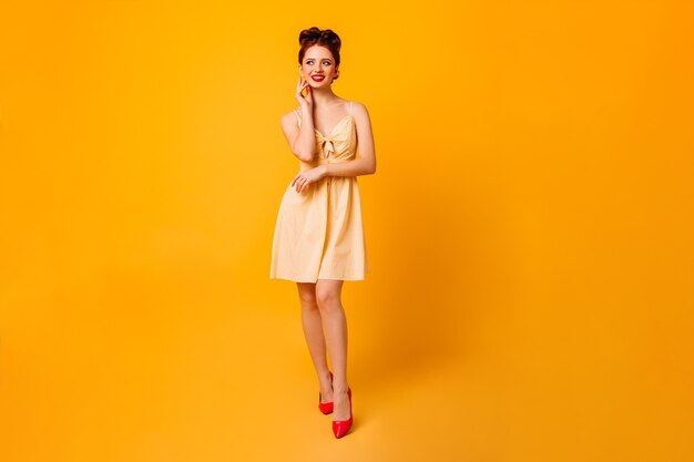 Vista de longitud completa de la alegre dama pinup. Mujer joven atractiva en vestido que se coloca en el espacio amarillo.