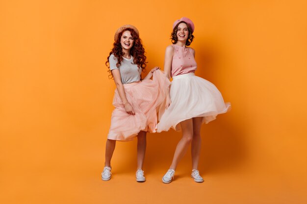 Vista de longitud completa de chicas entusiastas con cabello rizado bailando con una sonrisa. Foto de estudio de alegres amigas divirtiéndose sobre fondo amarillo.