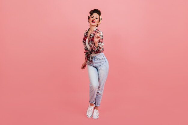 Vista de longitud completa de la impresionante dama pinup bailando sobre fondo rosa. Hermosa mujer morena en jeans y camisa a cuadros mirando a cámara.