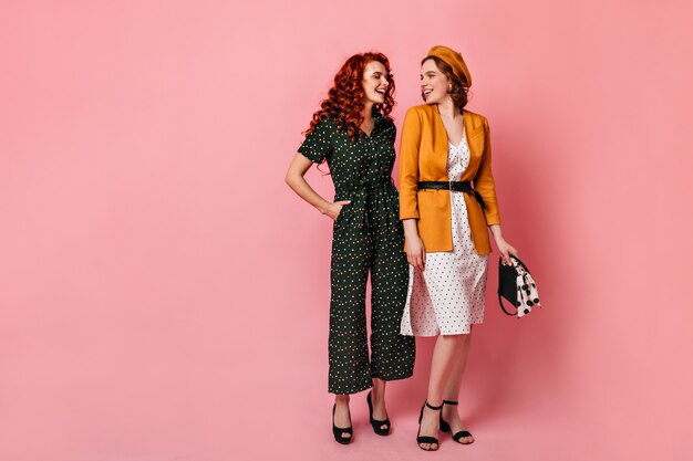 Vista de longitud completa de mujeres jóvenes en traje vintage. Foto de estudio de amigos alegres hablando sobre fondo rosa.