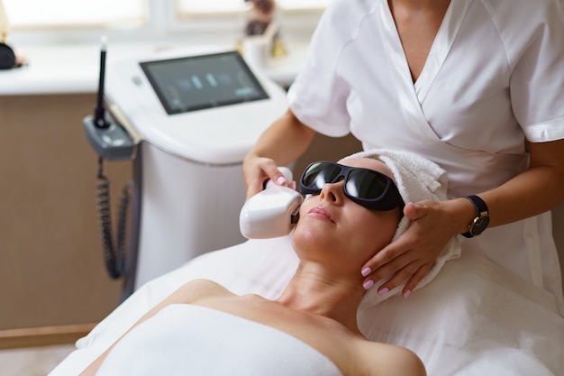 Vista del médico cosmetólogo haciendo un procedimiento anti-envejecimiento en la oficina de cosmetología. Mujer satisfecha con sombrero desechable acostado en el sofá y relajante.