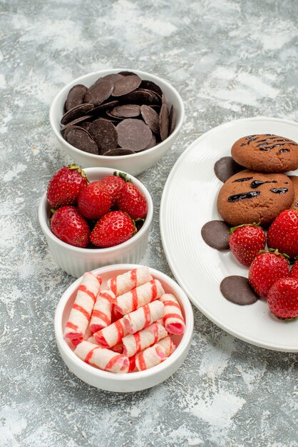 Foto gratuita vista de la mitad inferior de las galletas de chocolate, fresas y bombones redondos en la placa ovalada blanca y cuencos con caramelos, fresas, chocolates en el suelo gris-blanco