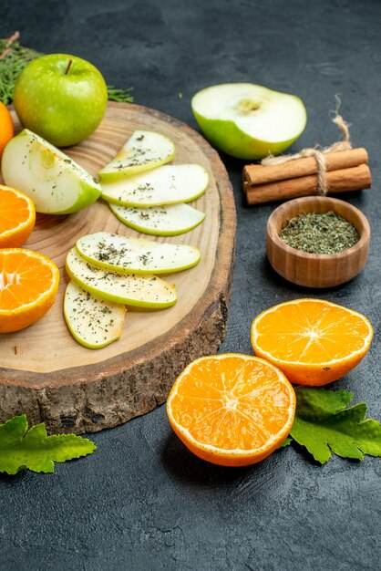 Vista de la mitad inferior rebanadas de manzana y naranja frescas sobre tablero de madera, polvo de menta seca en un tazón, ramas de canela, ramas de pino en la mesa negra