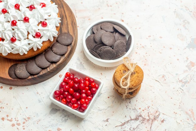 Vista de la mitad inferior de la torta con crema blanca en tazones de fuente de tabla de cortar con bayas y galletas de chocolate atadas con una cuerda en la mesa gris claro