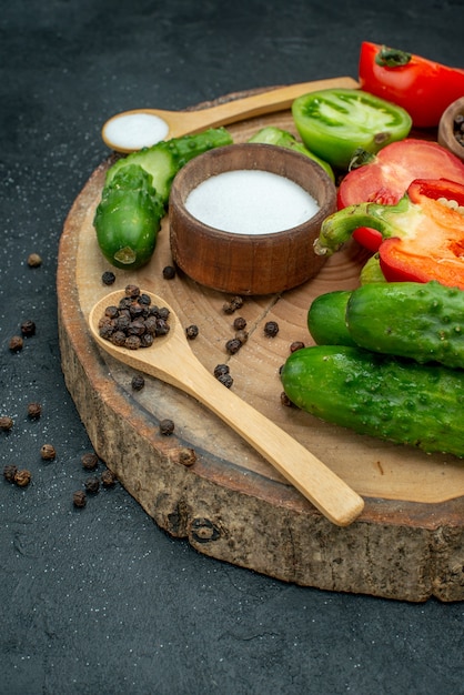 Vista de la mitad inferior verduras frescas pepinos pimienta negra y sal en cucharas de madera y tazones de fuente tomates rojos y verdes pimiento sobre tablero de madera en la mesa oscura