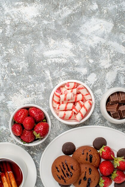 Vista de la mitad superior galletas fresas y chocolates redondos en el plato ovalado redondeado con cuencos de dulces fresas chocolates té de canela en la parte inferior de la mesa blanco grisáceo