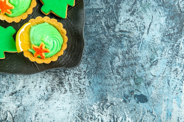 Vista de la mitad superior tartas pequeñas con crema pastelera verde galletas de árbol de Navidad en placa negra sobre superficie gris con espacio de copia