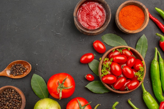 Vista de la mitad superior un tazón de tomates cherry pimientos rojos y verdes calientes y tomates hojas de laurel especias en cucharas de madera tazones de salsa de tomate pimiento rojo en polvo y pimienta negra en el suelo