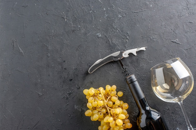 Vista de la mitad superior uvas amarillas frescas abridor de vino copa de vino y botella en mesa negra con espacio libre