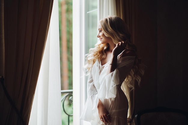 Vista de una mujer hermosa parada cerca del balcón y posando en lencería de encaje blanco. Chica en una habitación interior vintage, mirando por una ventana grande. Novia por la mañana.