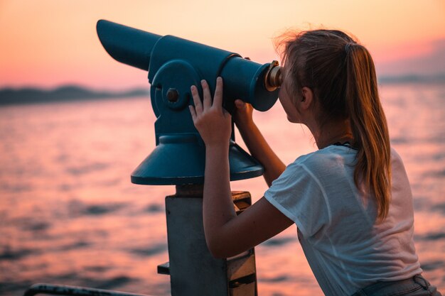 Vista de una mujer usando un telescopio y mirando hacia la puesta de sol en la playa desde el muelle