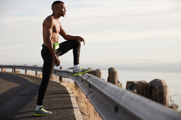 Vista panorámica de un joven físicamente activo determinado se encuentra en topless en la carretera, levanta la pierna en la señal de tráfico, hace ejercicios por la mañana, admira la naturaleza, le gusta el deporte, se siente exhausto, necesita energía