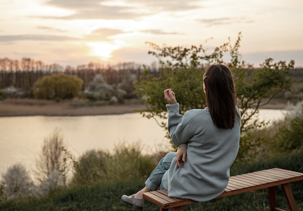 Foto gratuita la vista desde la parte de atrás de la niña mira la puesta de sol, sentada en un banco.