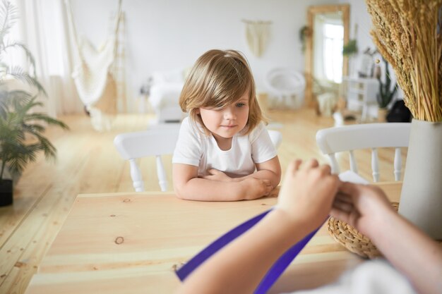 Vista desde la parte posterior de las manos de un niño irreconocible sosteniendo una hoja de papel violeta. Filmación en interiores de una linda niña con camiseta blanca sentada en el escritorio viendo a su hermano mayor hacer origami. Enfoque selectivo