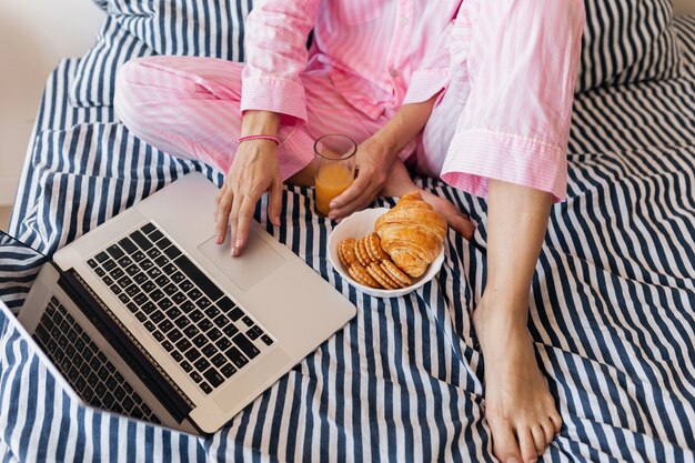 Vista desde la parte superior de la joven mujer bonita rubia en pijama rosa sentada en la cama con el portátil desayunando