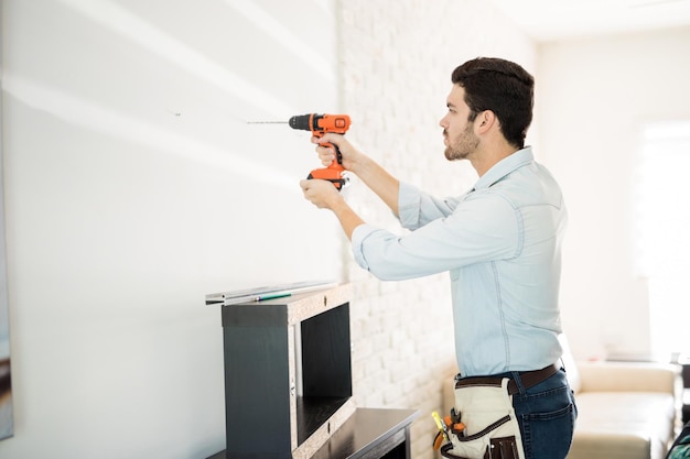 Vista de perfil de un apuesto joven manitas haciendo algunos agujeros en una pared e instalando un estante