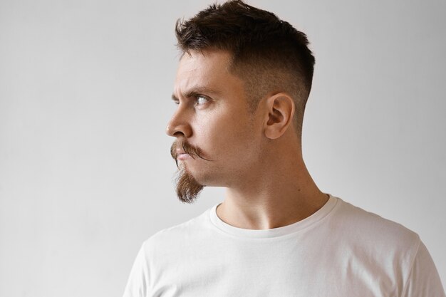 Vista de perfil de feroz y disgustado joven varón caucásico con perilla, bigote y elegante corte de pelo aplomo aislado en camiseta blanca mirando a otro lado con expresión de enojo ofendido, no quiere hablar