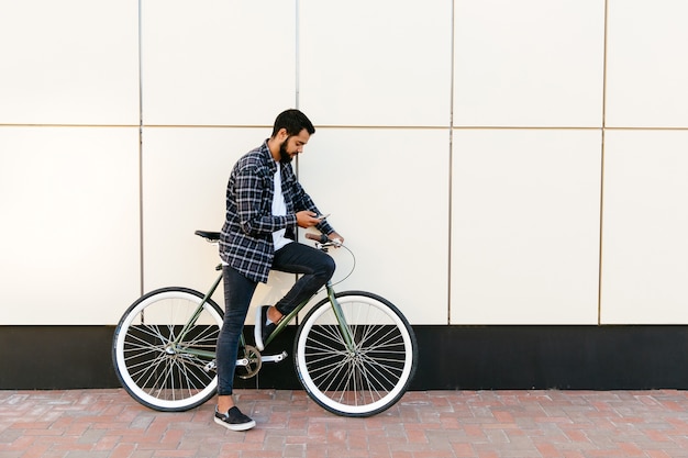 Vista de perfil de un hombre barbudo con estilo que usa un teléfono móvil mientras que se sienta en la bicicleta