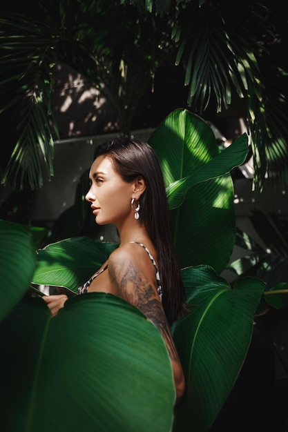 Vista de perfil de mujer morena caucásica con bronceado dorado usando bikini en vacaciones tropicales rodeada de hojas de palma tropicales en la selva