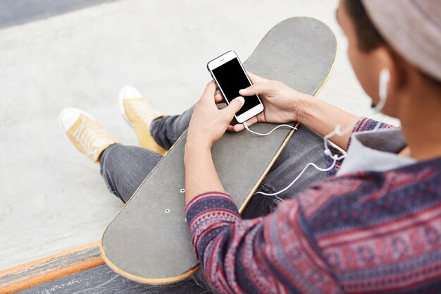 Vista posterior del adolescente hipster descansa en el parque de patinaje, patinetas con amigos, sostiene un teléfono inteligente con pantalla en blanco