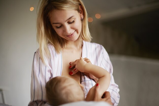 Vista posterior del adorable bebé de seis meses bebiendo leche materna. Atractiva joven europea en ropa de casa acunando a su hija en brazos, amamantandola, disfrutando de una conexión profunda