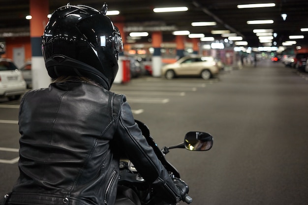 Vista posterior aislada de la motociclista conduciendo una moto deportiva de dos ruedas a lo largo del pasillo subterráneo del estacionamiento, yendo a estacionar su motocicleta después del paseo nocturno. Motociclismo, deportes extremos y estilo de vida