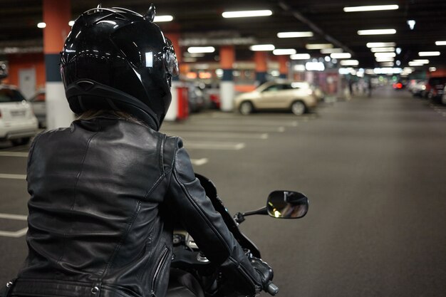 Vista posterior aislada de la motociclista conduciendo una moto deportiva de dos ruedas a lo largo del pasillo subterráneo del estacionamiento, yendo a estacionar su motocicleta después del paseo nocturno. Motociclismo, deportes extremos y estilo de vida