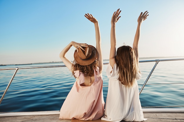 Vista posterior al aire libre de dos mujeres jóvenes en vacaciones de lujo, saludando a orilla del mar mientras está sentado en el yate.