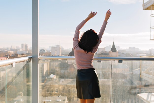 Vista posterior alegre joven atractiva que se extiende en la terraza en la soleada mañana en la vista de la gran ciudad. Mujer, gran éxito, felicidad, relajarse, humor alegre,