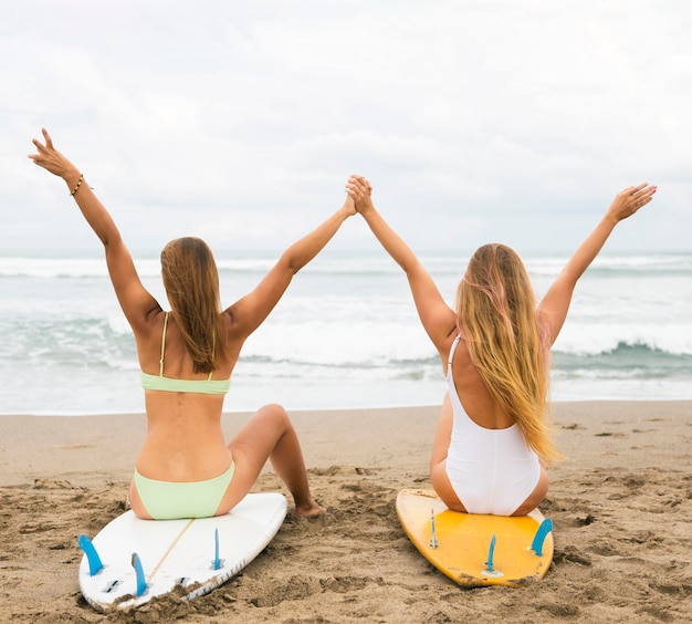 Vista posterior de amigas en la playa de pie sobre la tabla de surf con las manos arriba