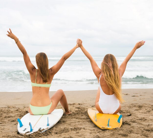 Vista posterior de amigas en la playa de pie sobre la tabla de surf con las manos arriba