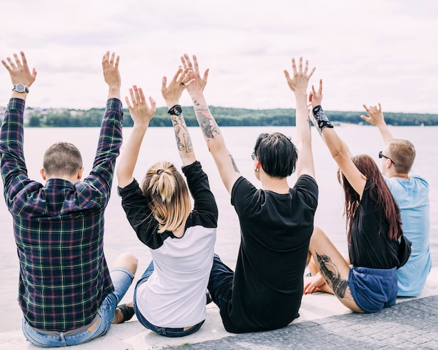 Vista posterior de amigos sentados cerca del lago levantando sus manos