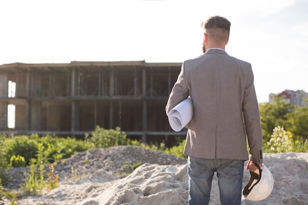 Vista posterior de la arquitectura masculina que sostiene el modelo y el casco que mira el emplazamiento de la obra