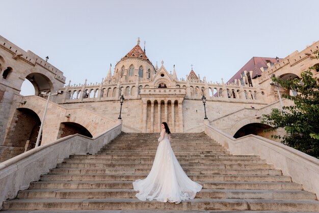 Vista posterior de una atractiva novia en las escaleras de un edificio histórico en la hermosa y cálida noche de verano
