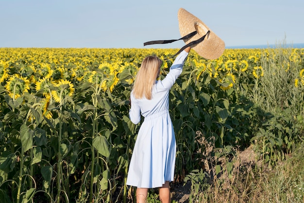Vista posterior chica sosteniendo su sombrero en un campo con flores de sol