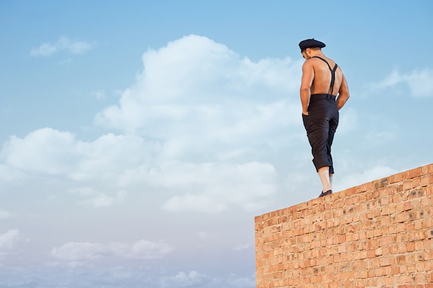 Vista posterior del constructor musculoso en ropa de trabajo de pie en la pared de ladrillo en lo alto. Hombre tomados de la mano en los bolsillos y mirando hacia abajo. Edificio extremo en un caluroso día de verano. Cielo azul de fondo.