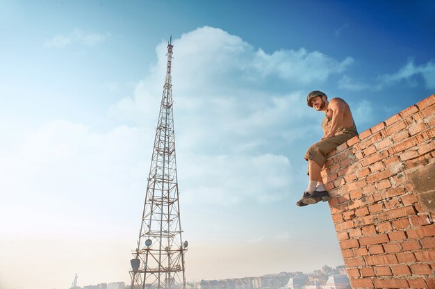 Vista posterior del constructor musculoso en ropa de trabajo de pie en la pared de ladrillo en lo alto. Hombre tomados de la mano en los bolsillos y mirando hacia abajo. Extrema en un caluroso día de verano. Cielo azul y alta torre de televisión en el fondo.