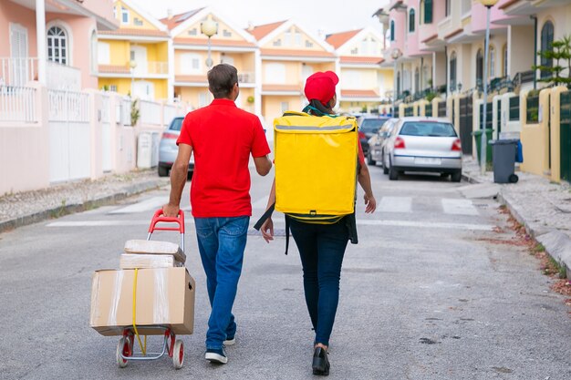 Vista posterior de dos mensajeros caminando con cajas en carro. Repartidores entregando el pedido en mochila térmica y vistiendo camiseta o gorra roja. Servicio de entrega y concepto de compra online.