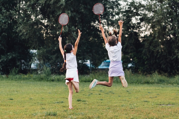 Vista posterior de dos niñas saltando en el parque con bádminton