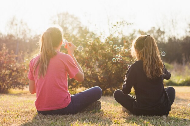 Vista posterior de dos niñas sentado en la hierba verde soplando burbujas