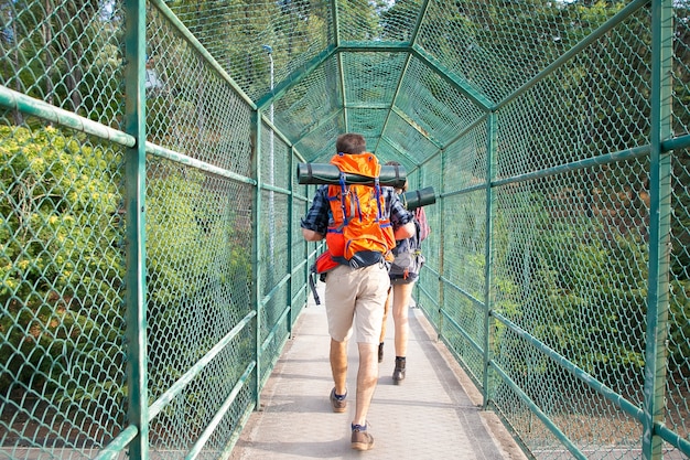 Vista posterior de los excursionistas caminando sobre el puente rodeado de rejilla verde. Dos turistas que llevan mochilas y atraviesan el camino. Concepto de turismo, aventura y vacaciones de verano.