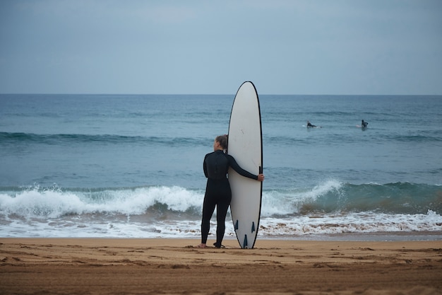 Vista posterior de la hermosa joven surfista abrazando su longboard en la orilla del océano y mirando las olas antes de surfear