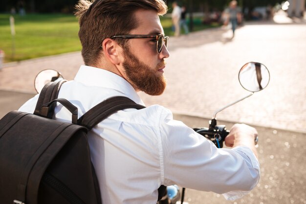 Vista posterior del hombre barbudo fresco en gafas de sol con mochila monta en moto moderna al aire libre y mirando a otro lado