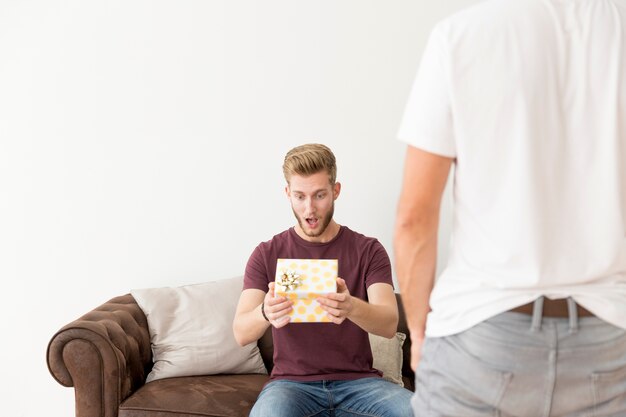 Vista posterior del hombre con el hombre sorprendido sentado en el sofá con la celebración de caja de regalo