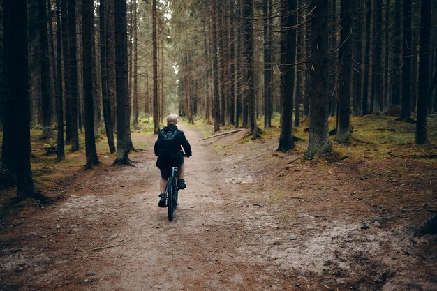 Vista posterior del hombre irreconocible montando bicicleta de montaña a lo largo de camino desierto en el bosque. Toma trasera de ciclismo masculino en el bosque en una mañana tranquila sin nadie alrededor. Concepto de personas, naturaleza y deportes.