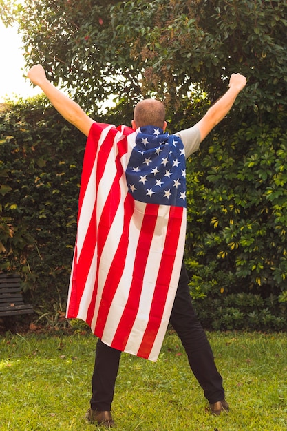 Vista posterior de un hombre de pie en el parque con la bandera de Estados Unidos capa levantando sus brazos