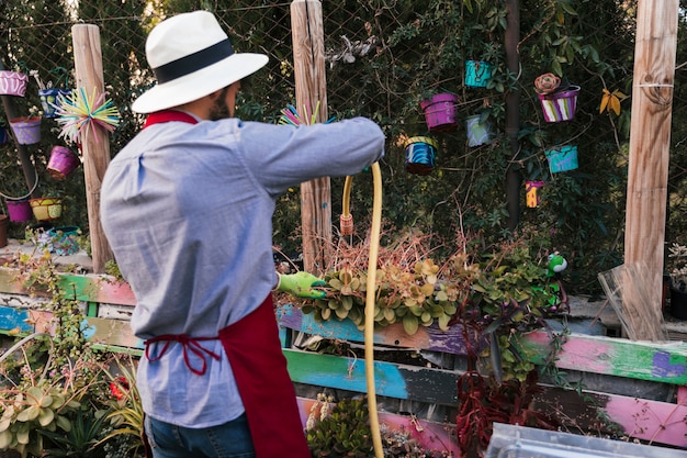 Vista posterior de un hombre con sombrero regando las plantas con manguera en el jardín