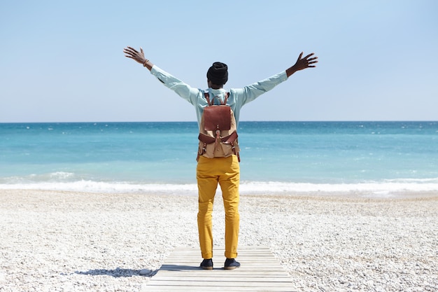 Vista posterior del irreconocible hombre europeo de piel oscura parado en el paseo marítimo de la playa tropical sintiéndose feliz y libre mientras ve el océano por primera vez durante el viaje de verano, manteniendo los brazos abiertos